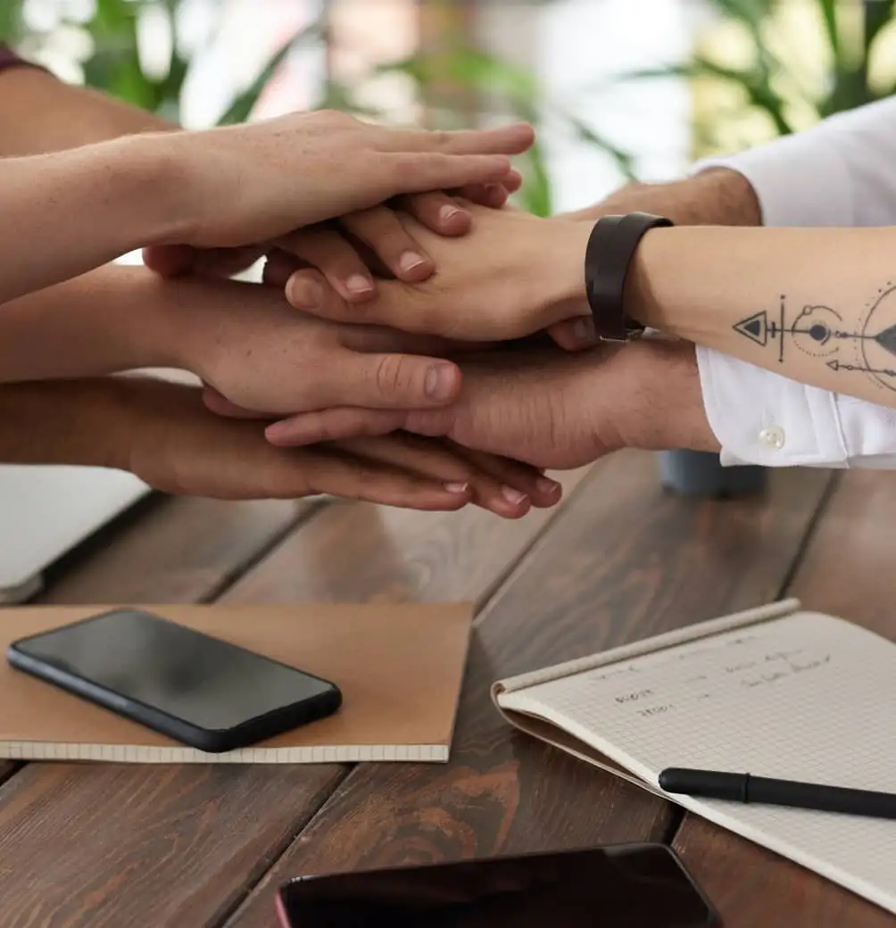 Hands stacked together on a wooden table beside a phone and a notebook