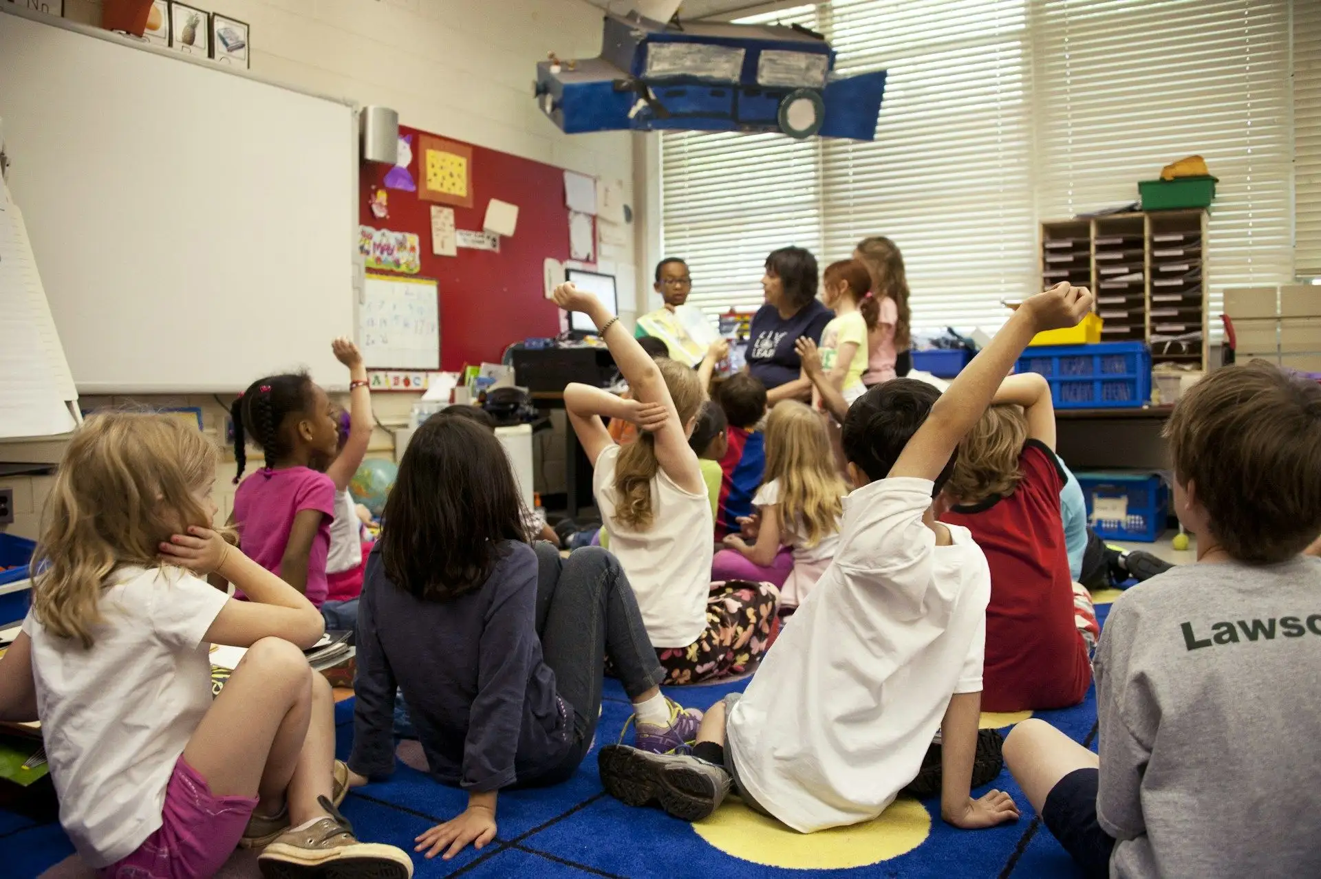 Children with hands raised on the carpet at a teacher's chair