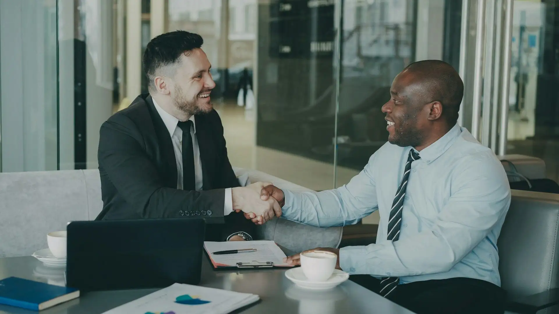 Two colleagues shaking hands across a table in a glass-walled office