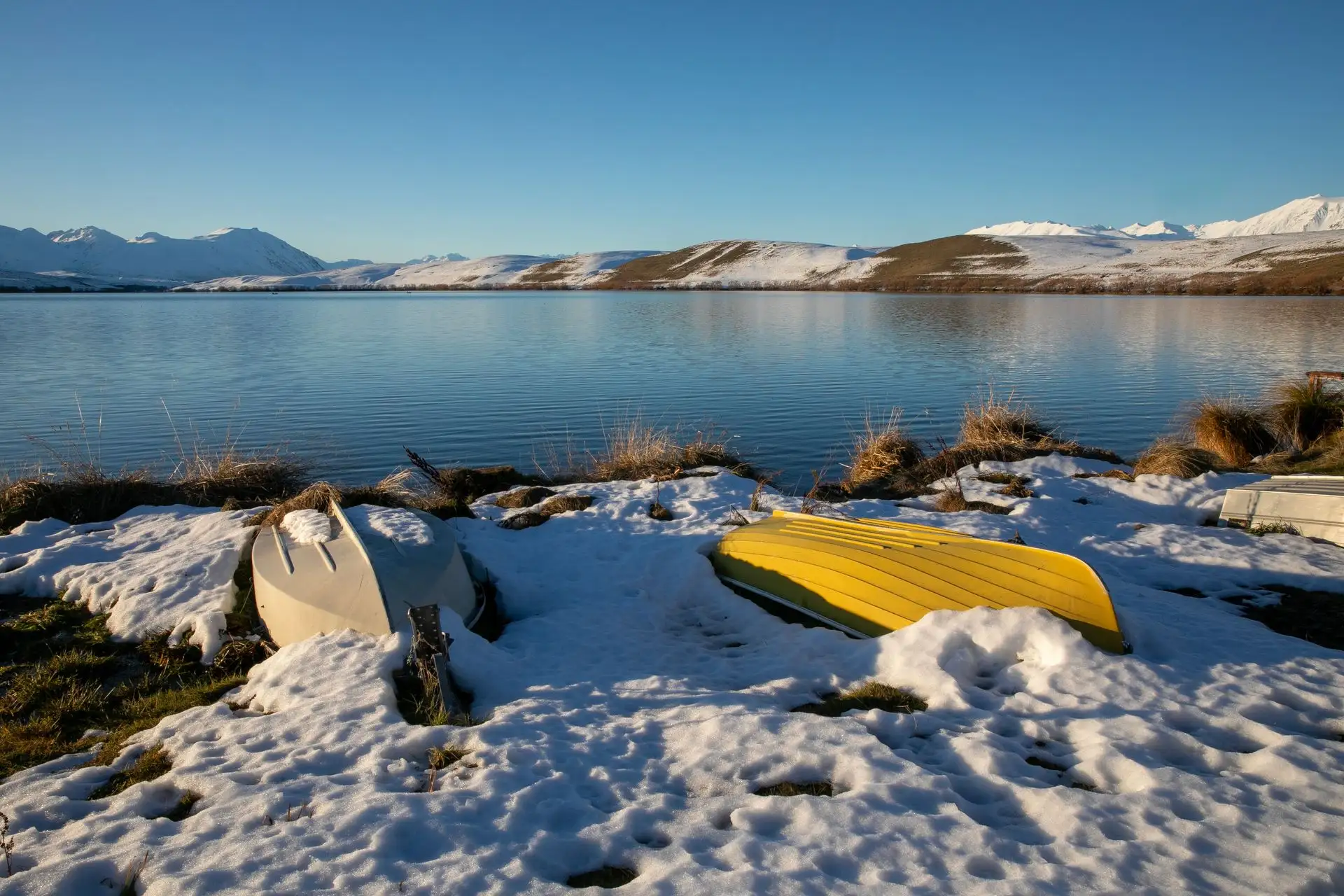 Upturned boats on snow beside a lake with snowy ranges behind