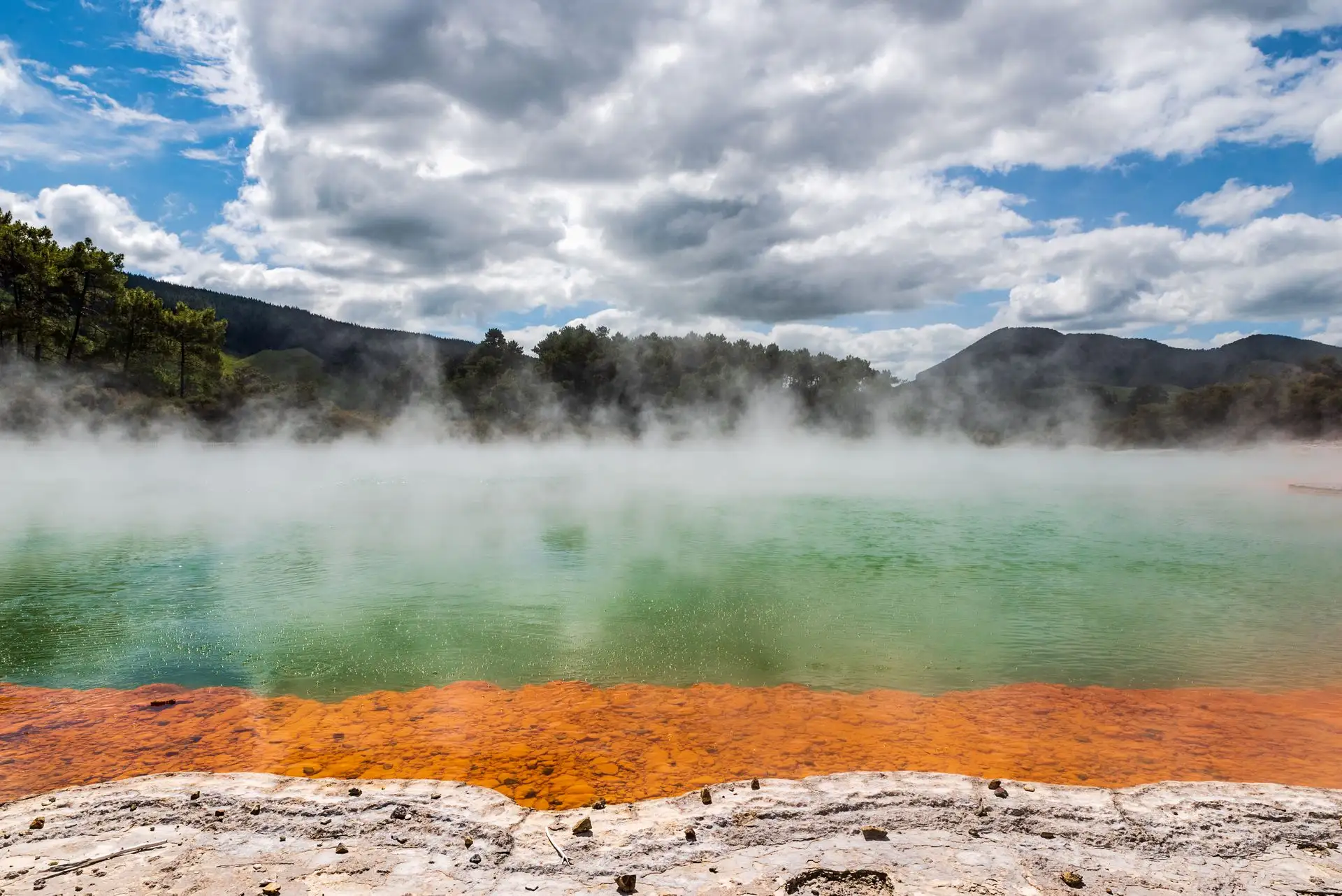 Mist over the orange-rimmed Champagne Pool at Wai-O-Tapu under a bright sky