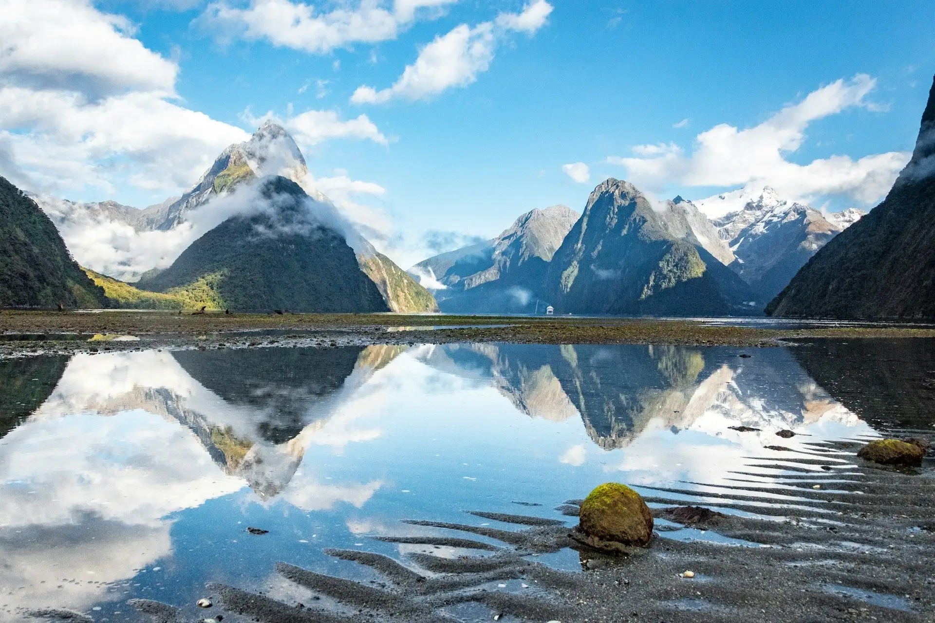 Mitre Peak and the snow peaks of Milford Sound mirrored in rippled shallows under a blue sky